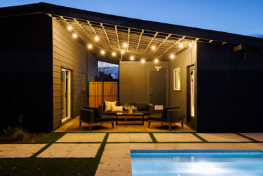 Modern backyard patio at dusk with string lights, covered lounge area featuring a cozy outdoor seating set and coffee table, and a lit swimming pool in the foreground