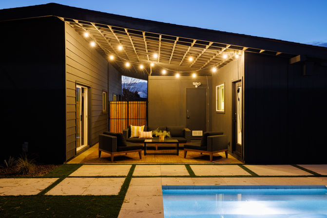 Modern backyard patio at dusk with string lights, covered lounge area featuring a cozy outdoor seating set and coffee table, and a lit swimming pool in the foreground
