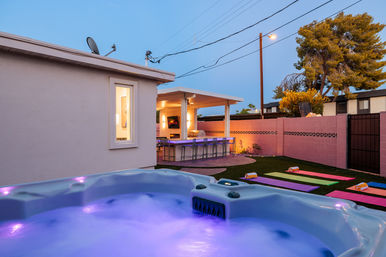 Evening suburban backyard patio with a glowing purple-lit hot tub in the foreground, covered outdoor bar and counter with stools, colorful yoga mats with rolled towels on the lawn, pink cinderblock wall and trees under a dusky sky