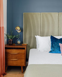 Cozy modern bedroom corner with tall olive upholstered headboard against teal wall, mid-century wooden nightstand, brass wall sconce, blue velvet pillow and coral curtains.