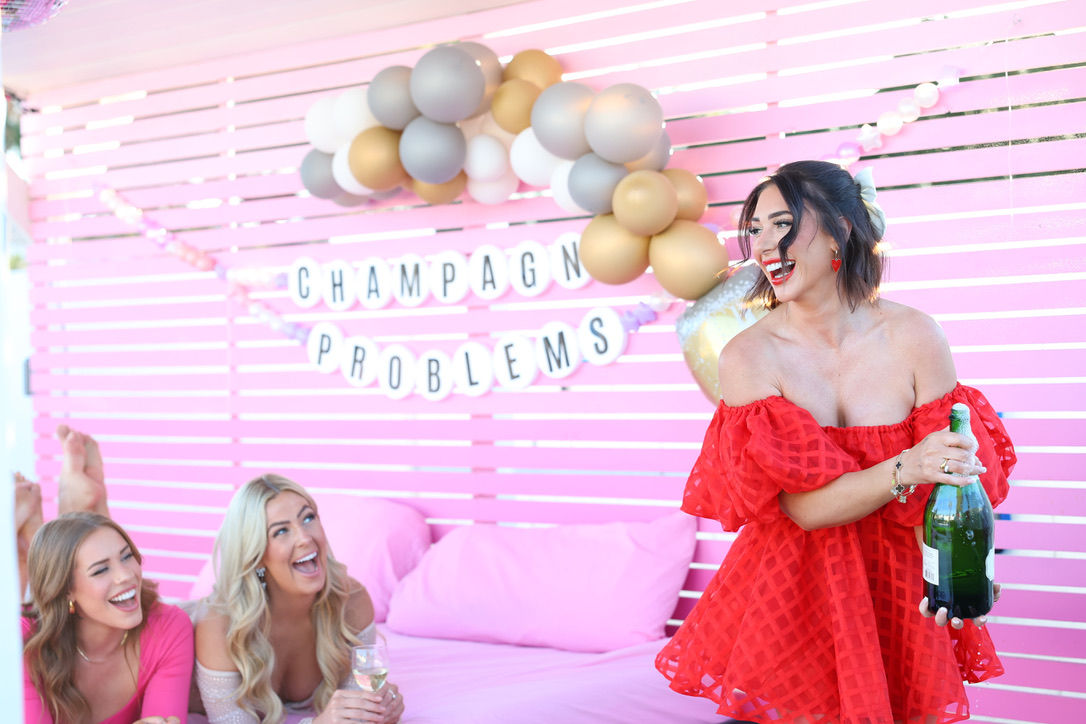 Three women at a pink party lounge with gold-and-gray balloons and a 'Champagne Problems' banner — one in a red dress popping a champagne bottle while two friends laugh on pink cushions.