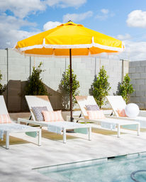 Sunlit backyard poolside patio with a bright yellow fringe umbrella shading white lounge chairs topped with pastel striped and polka-dot pillows beside a turquoise pool under a blue sky.