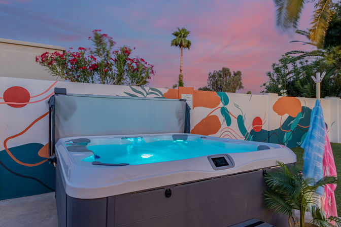 Glowing hot tub in a vibrant backyard at a pink sunset, colorful mural on the wall, palm tree, tropical plants and hanging towels.