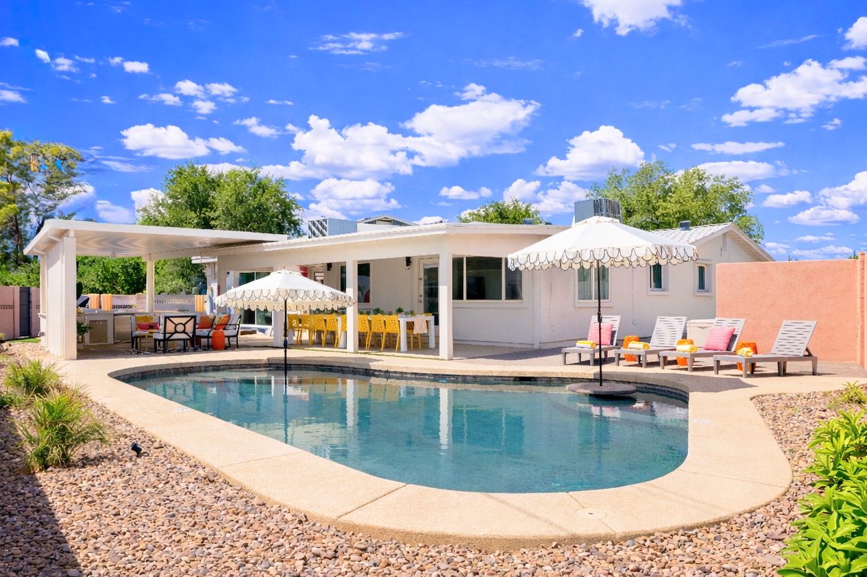Sunny suburban backyard with a kidney-shaped swimming pool, white scalloped umbrellas, lounge chairs with pink cushions, covered patio featuring yellow dining chairs and outdoor seating, gravel landscaping and bright blue sky with puffy clouds.