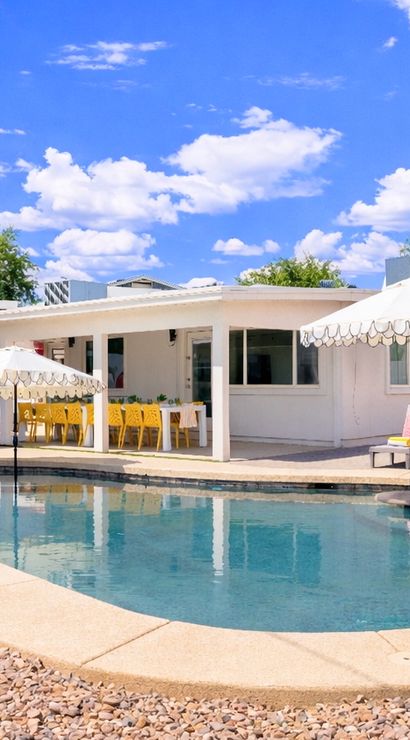 Sunny suburban backyard with a kidney-shaped swimming pool, white scalloped umbrellas, lounge chairs with pink cushions, covered patio featuring yellow dining chairs and outdoor seating, gravel landscaping and bright blue sky with puffy clouds.