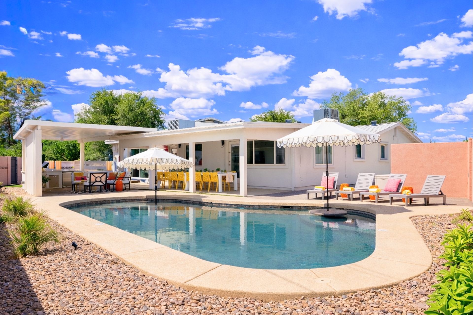 Sunny suburban backyard with a kidney-shaped swimming pool, white scalloped umbrellas, lounge chairs with pink cushions, covered patio featuring yellow dining chairs and outdoor seating, gravel landscaping and bright blue sky with puffy clouds.