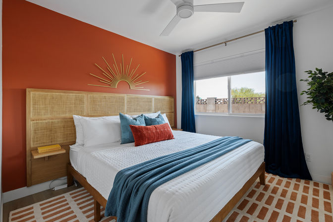 Modern bedroom with rust-orange accent wall and gold sunburst decor, woven rattan headboard, white quilted bed dressed with teal throw and rust-red lumbar pillow, navy curtains, large window, ceiling fan and patterned area rug