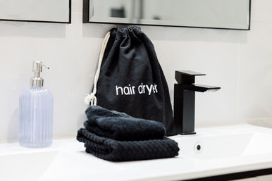 Modern white bathroom sink with matte black faucet, black drawstring bag labeled 'hair dryer', folded black towels, and a lilac glass soap dispenser.