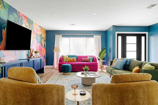 Colorful modern living room interior with bold floral accent wall, teal painted walls, fuchsia sofa, green velvet couch, mustard corduroy armchairs, blue media console with wall-mounted TV, round coffee table, wood floors, large window and black front door — bright eclectic home decor.