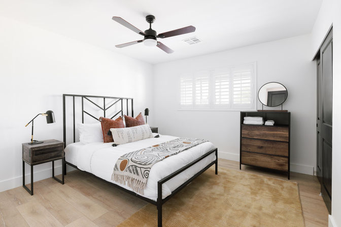 Bright modern bedroom with black metal bed frame, white bedding, patterned throw and rust accent pillows, wooden nightstand and dresser with round mirror, ceiling fan, plantation-shutter window, and light hardwood floors