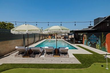 Sunny backyard with a long rectangular pool flanked by four wicker lounge chairs under fringed umbrellas, string lights overhead, a colorful muraled wall and a small putting green.