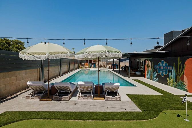 Sunny backyard with a long rectangular pool flanked by four wicker lounge chairs under fringed umbrellas, string lights overhead, a colorful muraled wall and a small putting green.