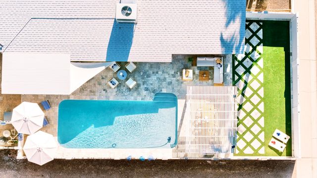 Aerial drone view of a modern backyard: turquoise rectangular pool, stone patio with umbrellas and lounge seating, pergola-covered dining area, and crisscross turf with cornhole boards.