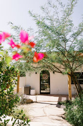 Sunny Southwestern-style desert courtyard with flagstone path to an arched stucco doorway topped with a wreath, shaded by a leafy tree and framed by blurred pink bougainvillea in the foreground.