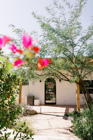 Sunny Southwestern-style desert courtyard with flagstone path to an arched stucco doorway topped with a wreath, shaded by a leafy tree and framed by blurred pink bougainvillea in the foreground.
