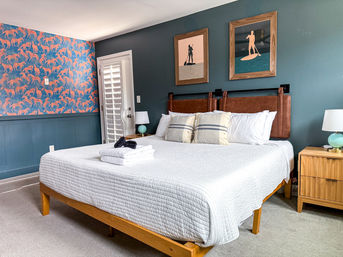 Coastal-inspired bedroom with a king bed dressed in a white quilt and folded towels, teal accent wall with two framed paddleboard prints, wooden headboard and nightstands with turquoise lamps, and vibrant orange animal-pattern wallpaper.