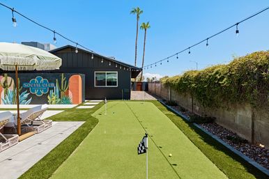 Sunny Scottsdale backyard with artificial putting green and checkered-flag pins, lounge chairs under a fringed umbrella, colorful mural on a dark house, hanging string lights and tall palm trees against a clear blue sky.