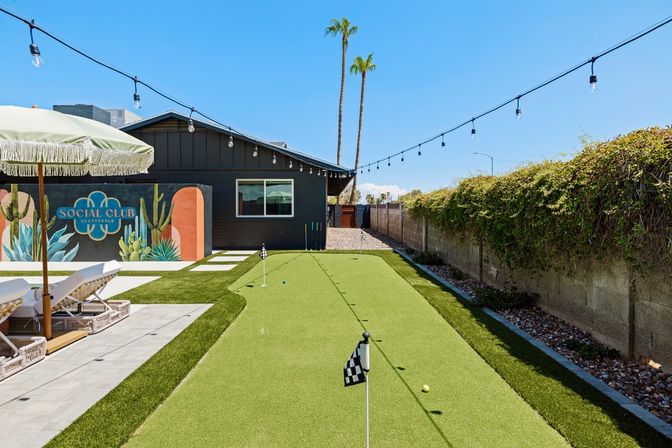 Sunny Scottsdale backyard with artificial putting green and checkered-flag pins, lounge chairs under a fringed umbrella, colorful mural on a dark house, hanging string lights and tall palm trees against a clear blue sky.