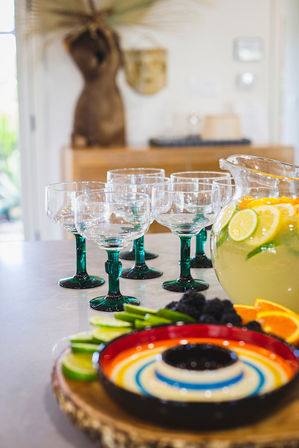 Green-stemmed cocktail glasses on a kitchen island next to a pitcher of citrus lemonade with lemon and lime slices and a colorful fruit platter on a wooden serving board — bright summer entertaining setup.