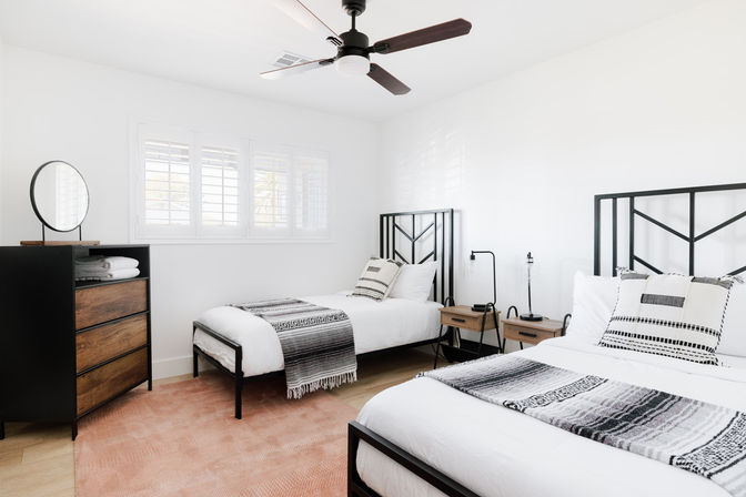 Cozy sunlit modern bedroom with two black metal-framed twin beds, white linens and gray patterned throws, wooden nightstands, dresser with round mirror, plantation-shuttered window and ceiling fan.