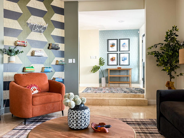 Cozy contemporary living room with a plush orange armchair, geometric accent wall with floating bookshelves, patterned rug, round wooden coffee table with decorative vase, indoor plants, and an entryway console with framed art.