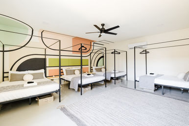 Bright modern shared dorm with multiple black four-poster single beds, colorful geometric wall mural, striped throws, large woven rug, ceiling fan and wall-mounted air conditioner.