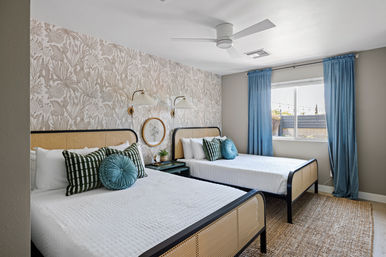 Bright coastal‑chic guest bedroom with two matching rattan headboard beds, botanical wallpaper accent wall, teal throw pillows and curtains, woven jute rug, modern ceiling fan, and sunlit window