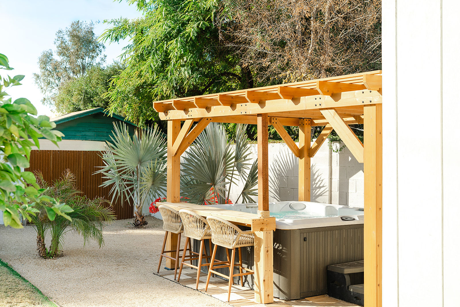 Sunny backyard patio with light-wood pergola over a hot tub and bar counter lined with woven rattan stools, surrounded by palm and silver fan palm plants and gravel landscaping.