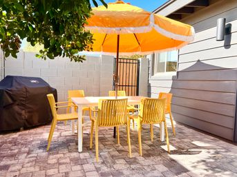 Cheerful sunny backyard patio with an orange fringed umbrella shading a white dining table surrounded by six yellow chairs on stone pavers, a covered grill, gray siding and cinderblock wall.