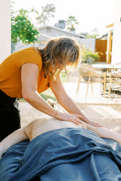 Massage therapist in an orange top giving a relaxing back massage to a client on a blue-sheeted table outdoors on a sunlit suburban patio.