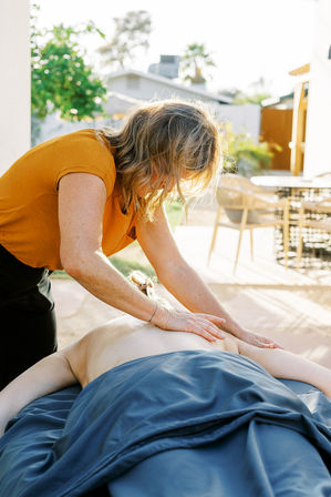 Massage therapist in an orange top giving a relaxing back massage to a client on a blue-sheeted table outdoors on a sunlit suburban patio.