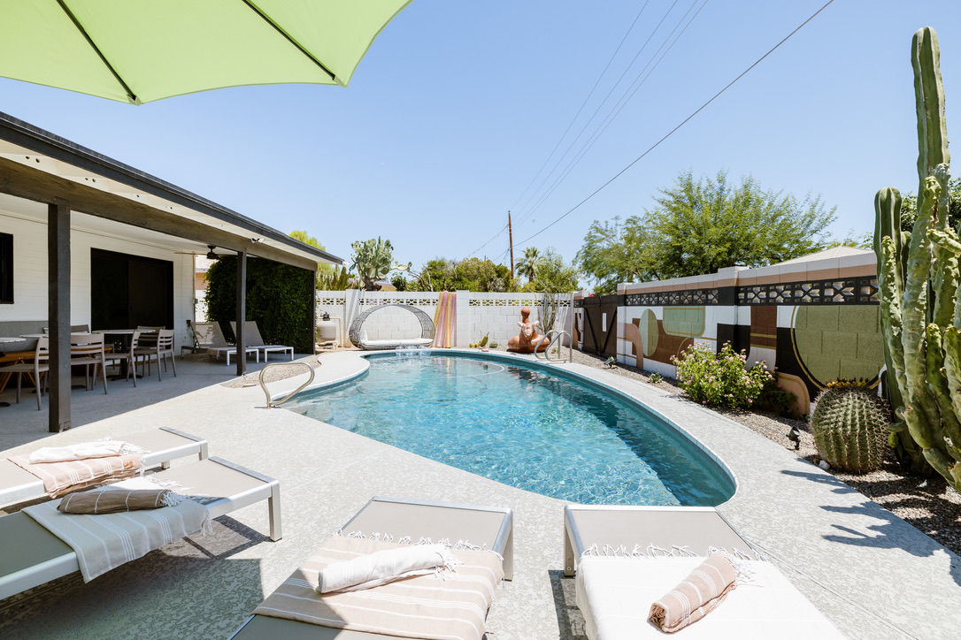 Sunny Southwestern backyard with kidney-shaped swimming pool, lounge chairs with towels, covered patio dining area, large cacti and mural-lined block wall