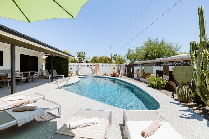 Sunny Southwestern backyard with kidney-shaped swimming pool, lounge chairs with towels, covered patio dining area, large cacti and mural-lined block wall
