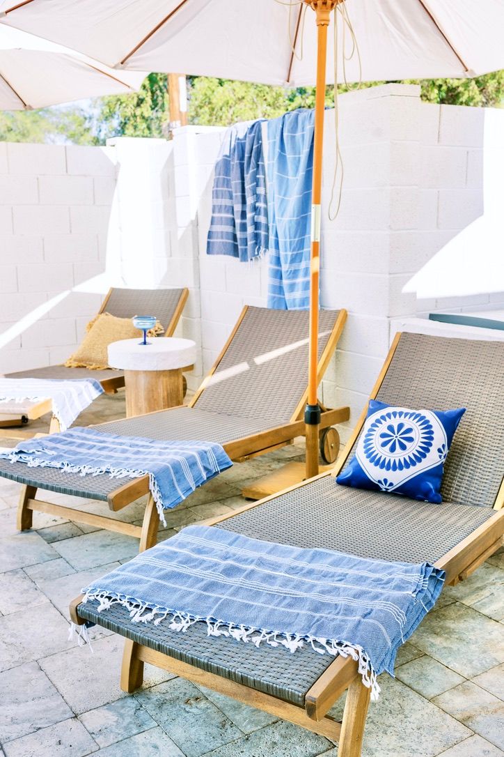 Sunlit outdoor patio with wooden sun loungers on stone tiles, blue-striped towels draped over the chairs, a blue patterned throw pillow on one lounger, and white umbrellas providing shade.