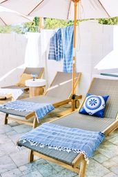 Sunlit outdoor patio with wooden sun loungers on stone tiles, blue-striped towels draped over the chairs, a blue patterned throw pillow on one lounger, and white umbrellas providing shade.