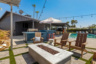 Sunlit modern backyard poolside patio with Adirondack chairs around a rectangular stone fire pit, outdoor bar and grill with stools, striped umbrella, string lights and palm trees under a clear blue sky.
