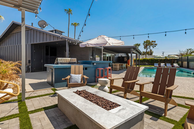 Sunlit modern backyard poolside patio with Adirondack chairs around a rectangular stone fire pit, outdoor bar and grill with stools, striped umbrella, string lights and palm trees under a clear blue sky.