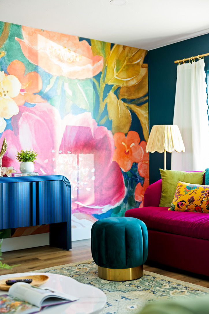Vibrant modern living room with a large watercolor floral mural, magenta velvet sofa with colorful throw pillows, teal velvet ottoman with gold base, blue ribbed console, scalloped floor lamp and patterned rug.