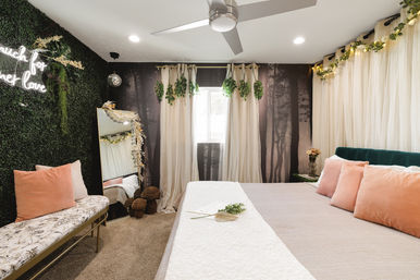 Bright modern boho bedroom interior with a forest mural and sheer curtains, velvet teal headboard, blush throw pillows, white bed runner, faux greenery wall with neon sign, hanging plants, full-length mirror and bench.