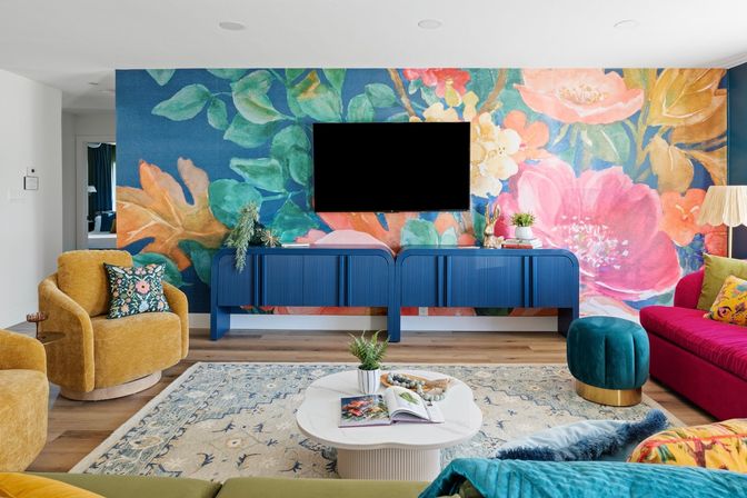 Vibrant modern living room with a large colorful floral mural, wall-mounted TV above a blue console, mustard velvet armchair, fuchsia sofa, teal ottoman, white coffee table on a patterned rug and hardwood floors — contemporary eclectic interior.