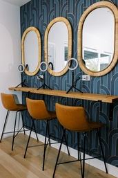 Three-station vanity with rattan oval mirrors, blue geometric wallpaper, floating wood shelf, tan leather bar stools and ring lights.