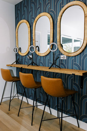 Three-station vanity with rattan oval mirrors, blue geometric wallpaper, floating wood shelf, tan leather bar stools and ring lights.