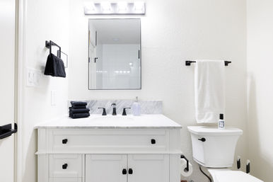 Clean bright contemporary bathroom with white marble vanity, black matte faucets and hardware, stacked dark hand towels, rectangular mirror and white towel over the toilet