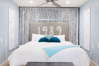Bright coastal-inspired bedroom featuring a large bed with white linens, gray headboard painted "1989", shimmering silver sequin backdrop, teal round accent pillows and an aqua throw, pale blue walls with bird prints and a ceiling fan.