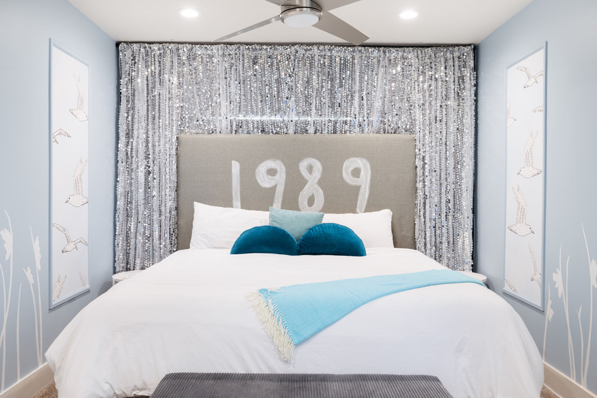 Bright coastal-inspired bedroom featuring a large bed with white linens, gray headboard painted "1989", shimmering silver sequin backdrop, teal round accent pillows and an aqua throw, pale blue walls with bird prints and a ceiling fan.
