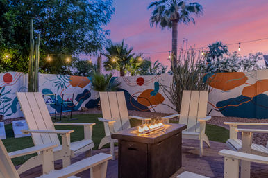 Cozy backyard patio at sunset with white Adirondack chairs around a lit rectangular fire pit, string lights overhead, palm trees and a colorful mural on the fence.