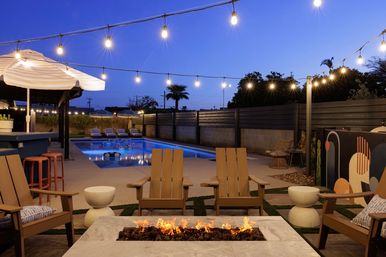 Cozy modern backyard patio at dusk with a lit rectangular swimming pool, poolside loungers, umbrellas, Adirondack chairs around a long gas fire pit, and string lights overhead.