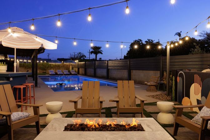 Cozy modern backyard patio at dusk with a lit rectangular swimming pool, poolside loungers, umbrellas, Adirondack chairs around a long gas fire pit, and string lights overhead.
