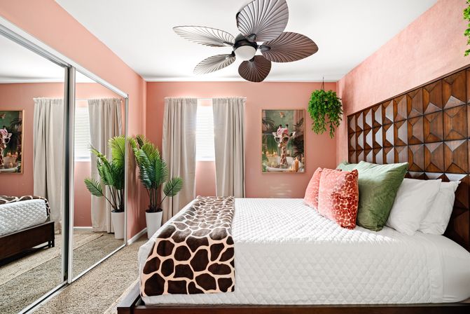 Sunlit coral bedroom interior with geometric wood headboard, white quilted bed topped with green and coral pillows and a giraffe-print throw, tropical potted plants, hanging fern, mirrored closet doors and oversized leaf-style ceiling fan.