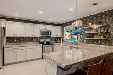 Sunny kitchen with white cabinets, large granite island, dark botanical wallpaper, stainless appliances, pendant lights, open shelves and a poolside window view.